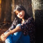 Un retrato sereno de una mujer joven sentada en un bosque otoñal iluminado por el sol, capturando la tranquilidad de la naturaleza.