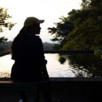 Silueta de una mujer sentada junto a una fuente de agua reflectante rodeada de árboles al atardecer, creando una atmósfera tranquila.