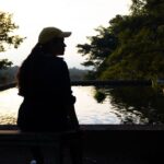 Silueta de una mujer sentada junto a una fuente de agua reflectante rodeada de árboles al atardecer, creando una atmósfera tranquila.