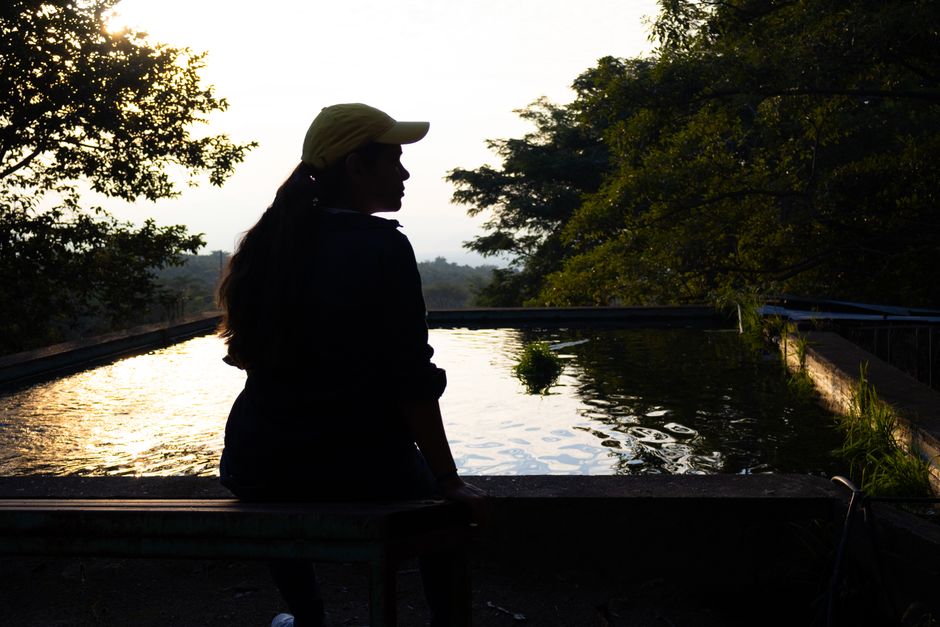 Silueta de una mujer sentada junto a una fuente de agua reflectante rodeada de árboles al atardecer, creando una atmósfera tranquila.
